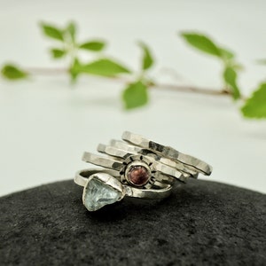 May include: A set of five silver rings with hammered textures. One ring features a pink gemstone, and another ring features a light blue gemstone.