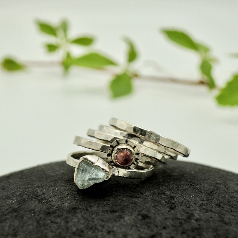 May include: A set of five silver rings with hammered textures. One ring features a pink gemstone, and another ring features a light blue gemstone.
