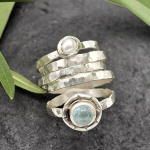 May include: A set of four silver rings with hammered textures. The top ring features a white pearl, and the bottom ring has a light blue gemstone.