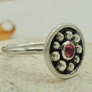 May include: A silver ring with a round, textured bezel set with a single red gemstone. The bezel is surrounded by a circle of small silver beads.