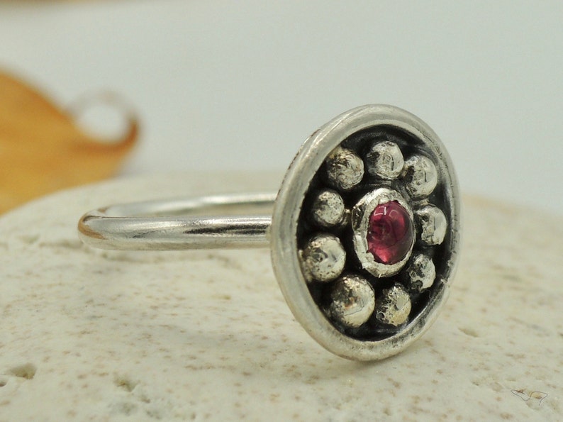 May include: A silver ring with a round, textured bezel set with a single red gemstone. The bezel is surrounded by a circle of small silver beads.