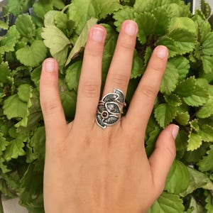 May include: A silver ring with a design of overlapping leaves and a red gemstone in the center.