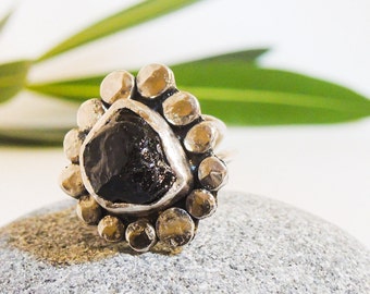 May include: A silver ring with a large, irregularly shaped black stone at the center, surrounded by smaller, textured silver beads. The ring is set on a gray stone surface, with a blurred green leaf in the background.