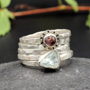 May include: A silver ring set with a light blue aquamarine and a dark pink gemstone. The ring is made of four bands, each with a hammered texture.