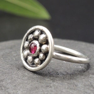 May include: A silver nose ring with a round, textured design featuring a small, red gemstone in the center.