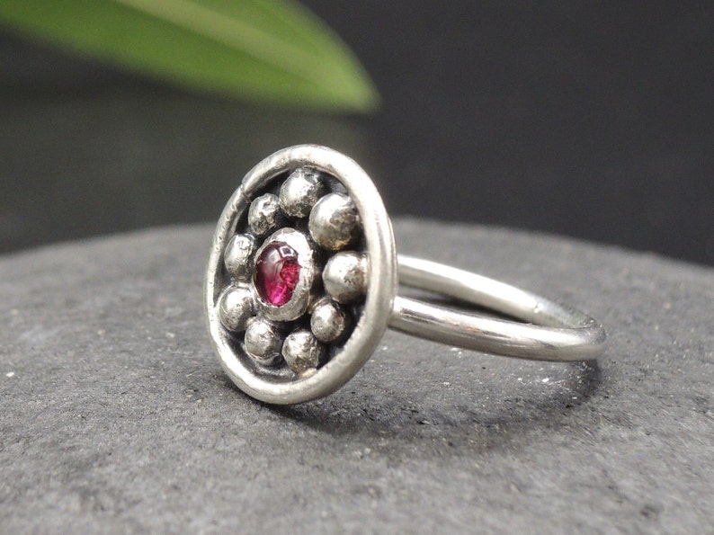 May include: A silver nose ring with a round, textured design featuring a small, red gemstone in the center.