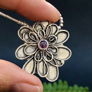Può includere: Una collana con un ciondolo a forma di fiore d'argento con una pietra preziosa viola al centro. Il ciondolo è dettagliato con intricati vortici e trame.