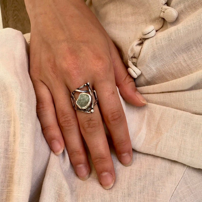 May include: A silver ring with a geometric design featuring a large, irregular, light green stone.