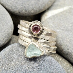 May include: A set of five silver rings with hammered textures. One ring features a pink gemstone set in a circular bezel with a decorative border. Another ring features a light blue gemstone.