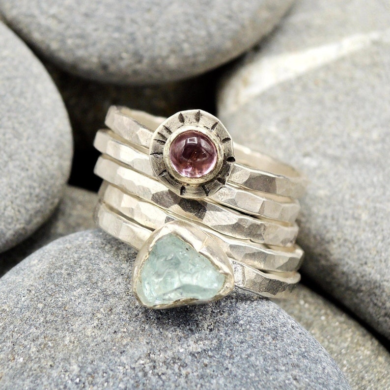May include: A set of five silver rings with hammered textures. One ring features a pink gemstone set in a circular bezel with a decorative border. Another ring features a light blue gemstone.