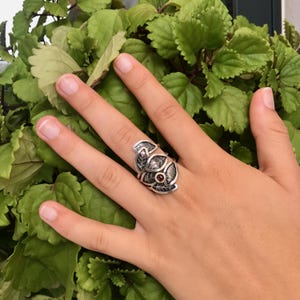 May include: A silver ring with a design of leaves and a red gemstone in the center.