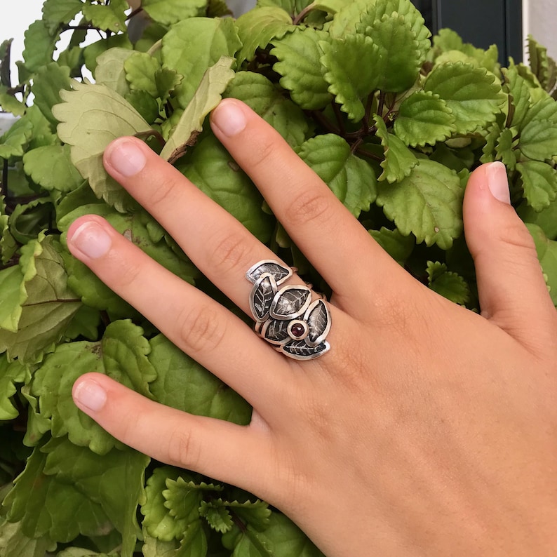 May include: A silver ring with a design of leaves and a red gemstone in the center.