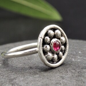 May include: A silver nose ring with a round, textured design featuring a small pink gemstone in the center.