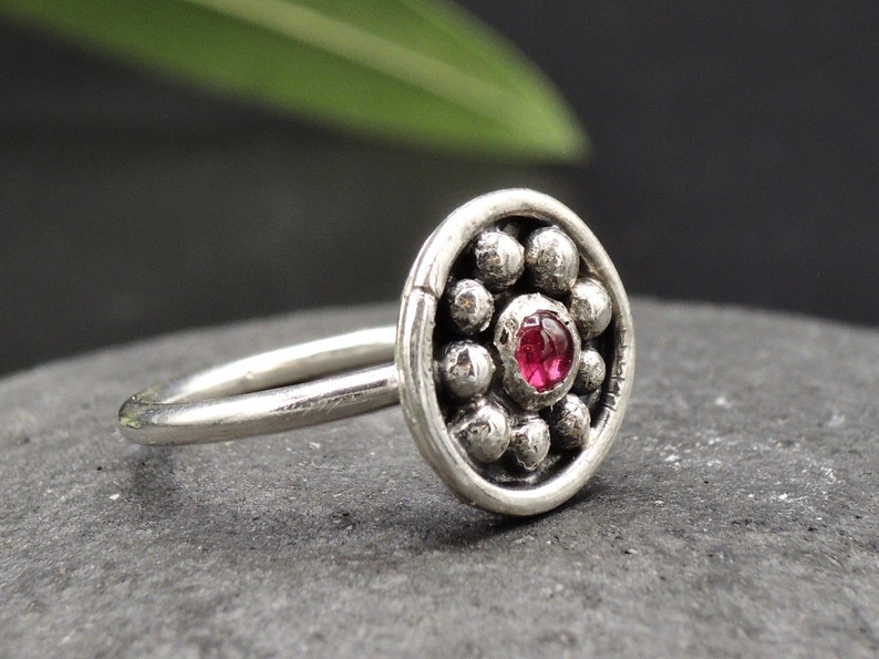 May include: A silver nose ring with a round, textured design featuring a small pink gemstone in the center.