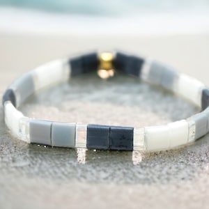 May include: A gray, white, and black beaded bracelet with a gold clasp. The bracelet is on a white sandy surface.