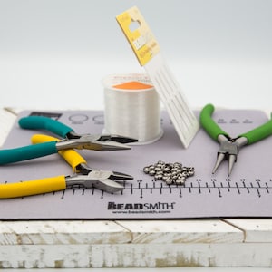May include: A gray beading mat with a ruler, a spool of clear elastic cord, two pairs of pliers with yellow and green handles, and a pile of silver beads.