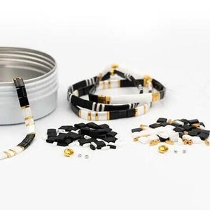 May include: A set of black, white, and gold beads in a round tin container. The beads are arranged in a circle and are ready to be strung together to make a bracelet. The tin container has the words "Make & Share" printed on the lid.