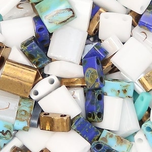 May include: A collection of two-hole beads in a circular container. The assortment includes solid white rectangular beads, metallic bronze rectangular beads, and multi-colored beads featuring blue, turquoise, and brown marbled patterns.