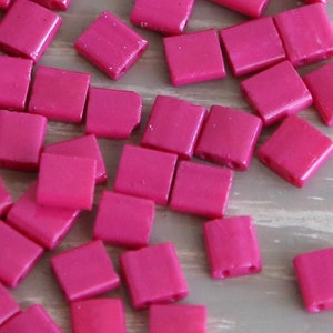 May include: A large pile of small, square, pink plastic beads. The beads have a glossy finish and are scattered on a light brown surface.