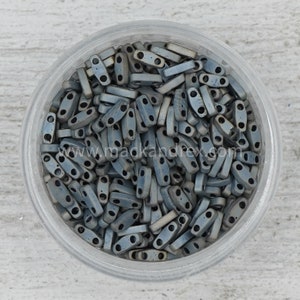 May include: A clear glass jar filled with a large quantity of small, gray, rectangular beads with two holes in each bead. The beads are arranged in a random pattern.