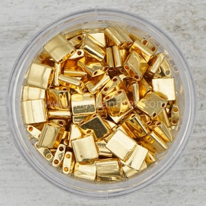 May include: A clear plastic container filled with gold-colored metal beads. The beads are small and have a rectangular shape.