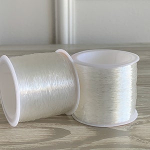 May include: Two spools of clear, elastic beading cord. The cord is wound around white plastic spools. The cord appears translucent and slightly iridescent. The spools are resting on a light-colored wooden surface.