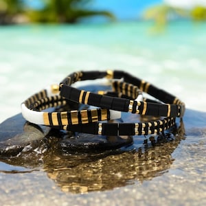 May include: Three stacked bracelets with black and gold beads. The bracelets are on a gray rock with a blurry background of blue water and green palm trees.