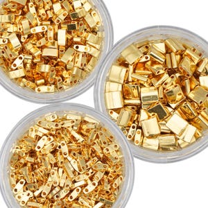 May include: Three clear containers filled with gold-colored metal beads. The beads are small and rectangular with rounded edges. The containers are on a white background.