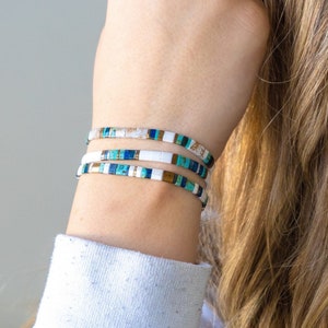 May include: A stack of four bracelets with white, blue, and gold beads. The bracelets are stacked on a person's wrist.
