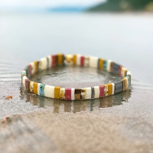May include: A colorful beaded bracelet with white, yellow, pink, blue, and gray beads. The bracelet is made of small, square-shaped beads.