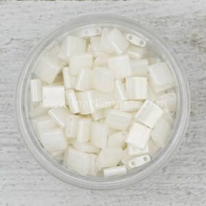 May include: A close-up of a clear glass jar filled with small, white, square-shaped beads. The beads have a slightly iridescent finish.