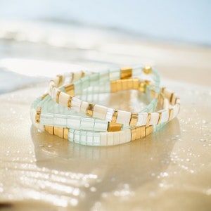 May include: A stack of three bracelets with light blue, white, and gold beads. The bracelets are made of square beads and are stacked on top of each other.