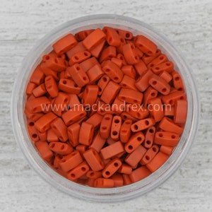 May include: A close-up of a jar filled with small, orange, rectangular beads. The beads have two holes in each. The jar is on a white surface. www.mackandrex.com