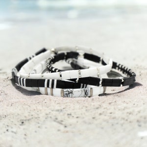 May include: A set of four bracelets made of black and white ceramic tiles. The bracelets are stacked on top of each other and are lying on a white sandy surface.