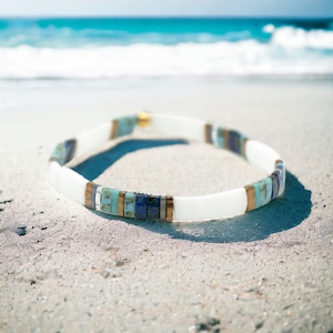 May include: A white beaded bracelet with blue, turquoise, and gold accents. The bracelet is lying on a sandy beach.