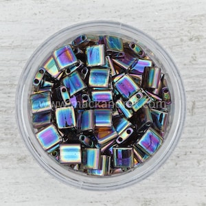 May include: A clear plastic container filled with iridescent, rectangular beads. The beads have a flat, square shape with two holes on one side. The beads reflect a rainbow of colors, including blue, purple, and green.