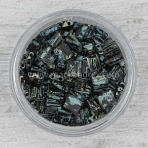 May include: A close-up view of a small container filled with black and teal square-shaped beads with white and yellow accents. The beads have a textured, marbled appearance.