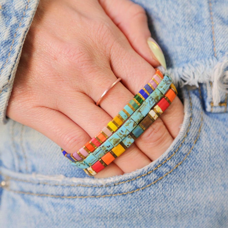 May include: A hand wearing a stack of colorful beaded bracelets with a gold ring on the finger. The bracelets are made of small square beads in various colors, including turquoise, red, orange, yellow, green, and blue.