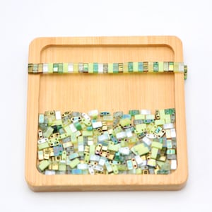 May include: A wooden tray filled with a variety of green, yellow, and silver glass beads. The beads are arranged in a random pattern and are all of the same size and shape. There is a single row of beads at the top of the tray.