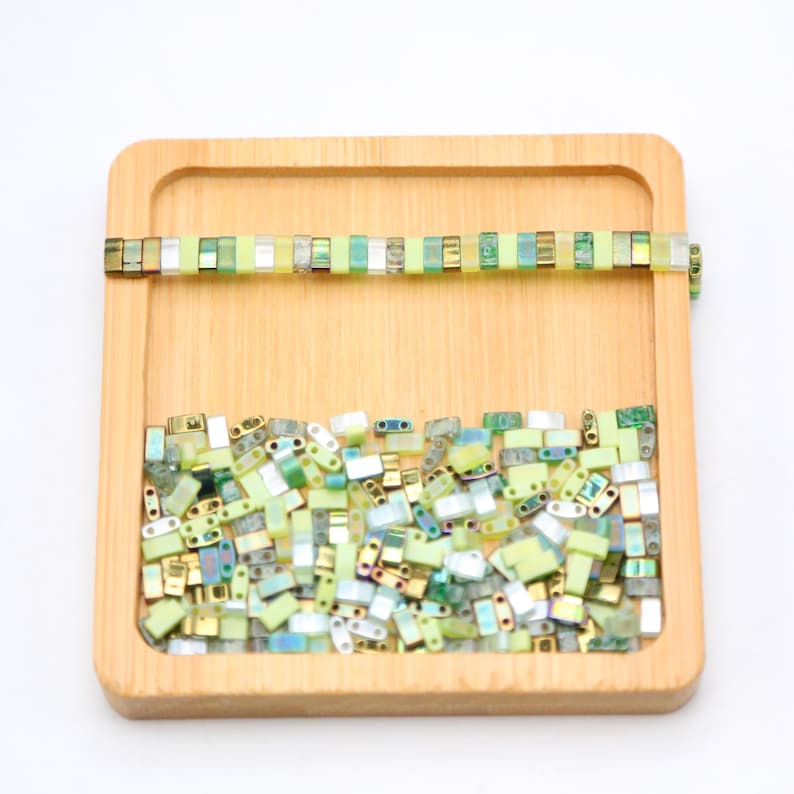 May include: A wooden tray filled with a variety of green, yellow, and silver glass beads. The beads are arranged in a random pattern and are all of the same size and shape. There is a single row of beads at the top of the tray.