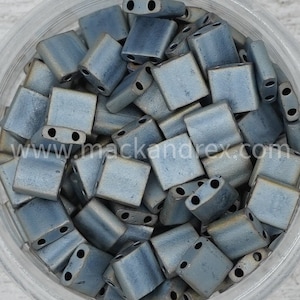 May include: A close-up of a container filled with small, square, gray-blue glass beads. The beads have two holes on each side and are designed for use in jewelry making.