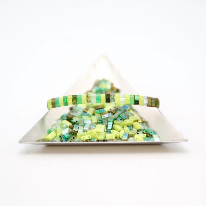 May include: A close-up of a green and yellow striped bracelet made of small square beads. The bracelet is sitting on a silver tray with a pile of loose green and yellow square beads.