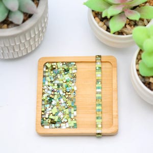 May include: A wooden tray with a pile of green and gold square beads and a strand of green and gold beads on the right side.
