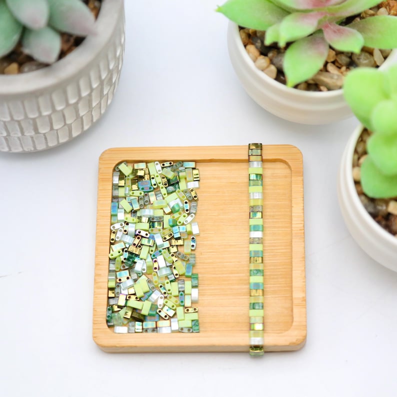 May include: A wooden tray with a pile of green and gold square beads and a strand of green and gold beads on the right side.