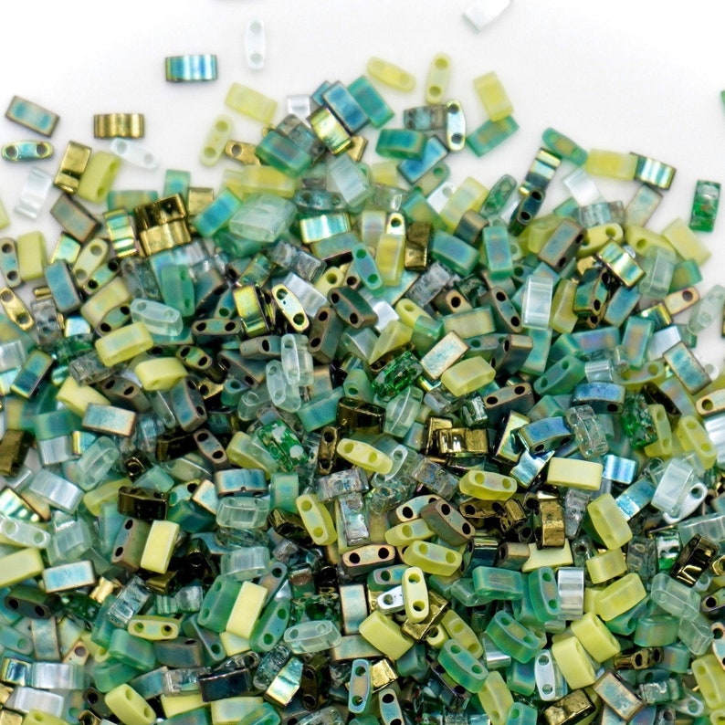 May include: A large assortment of green, yellow, and clear glass seed beads. The beads are all rectangular in shape and have two holes.
