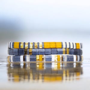 May include: Three stacked bracelets with a pattern of gray, white, and yellow beads.