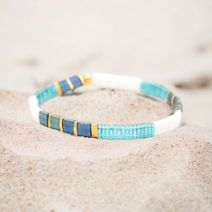 May include: A blue and white beaded bracelet with gold accents. The bracelet is made of small, square beads and is designed to look like a beach scene.