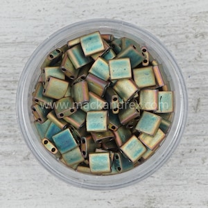 May include: A close-up of a clear plastic container filled with small, square, iridescent green and gold beads. The beads have two holes in each. The container is sitting on a light wood surface.