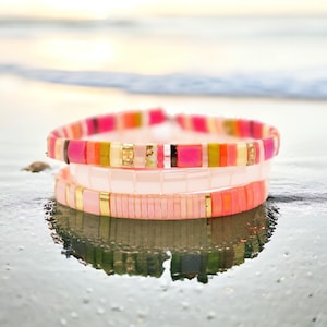 May include: Three stacked bracelets with pink, orange, yellow, and white beads. The bracelets are made of small, square beads and have a subtle shimmer.