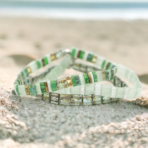 May include: Three stacked bracelets made of green, white, and gold glass beads. The bracelets are on a sandy surface.